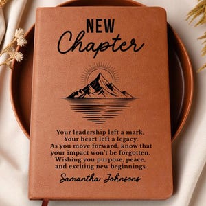 May include: A brown leather-bound journal with the words "NEW Chapter" and a mountain illustration. The journal includes a personalized message and the name "Samantha Johnsons" at the bottom. The journal is resting on a brown plate.