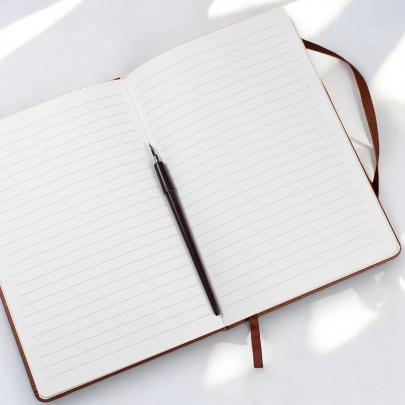 May include: An open lined notebook with a brown pen lying on the open pages. The notebook has a brown leather cover and a brown ribbon bookmark.
