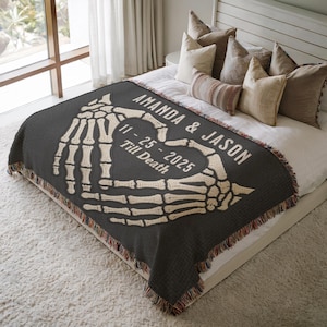 May include: A dark gray throw blanket with a cream-colored skeleton hand design forming a heart. The blanket has the names "AMANDA & JASON" and the date "11-25-2025" with the words "Till Death" printed on it. The blanket has a fringed edge.