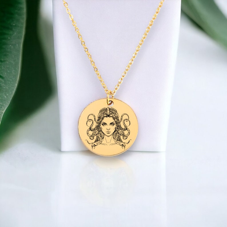 May include: A gold necklace with a round pendant featuring a black and white illustration of Medusa, a Greek mythological figure with snakes for hair.