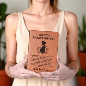 May include: A brown leather journal with the text "TO MY SISTER A BEAUTIFUL MOM-TO-BE" and a silhouette of a pregnant woman. The journal also includes a heartfelt message and the signature "With love, Amanda."