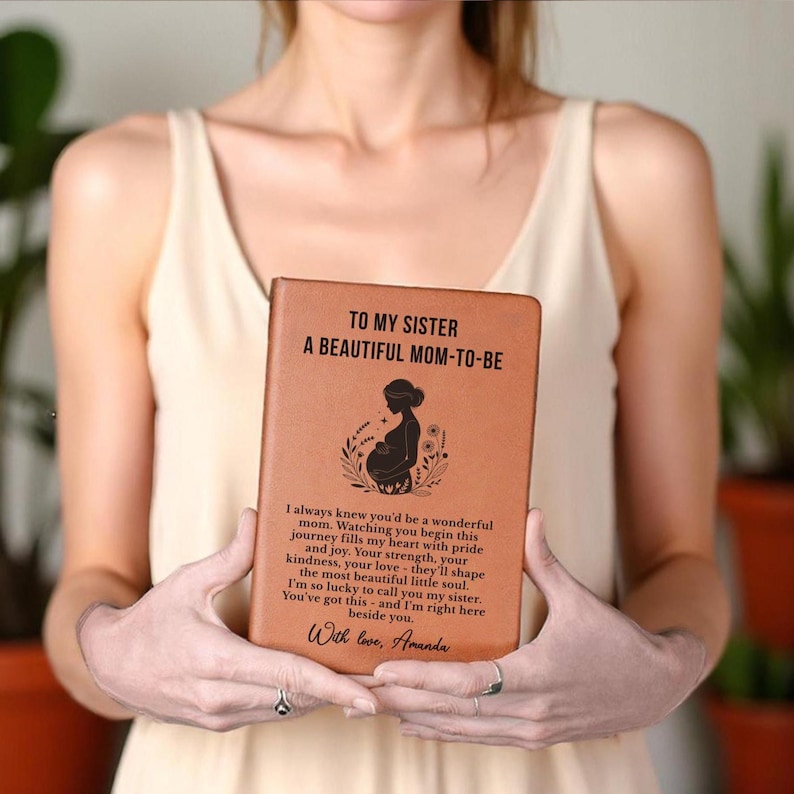 May include: A brown leather journal with the text "TO MY SISTER A BEAUTIFUL MOM-TO-BE" and a silhouette of a pregnant woman. The journal also includes a heartfelt message and the signature "With love, Amanda."