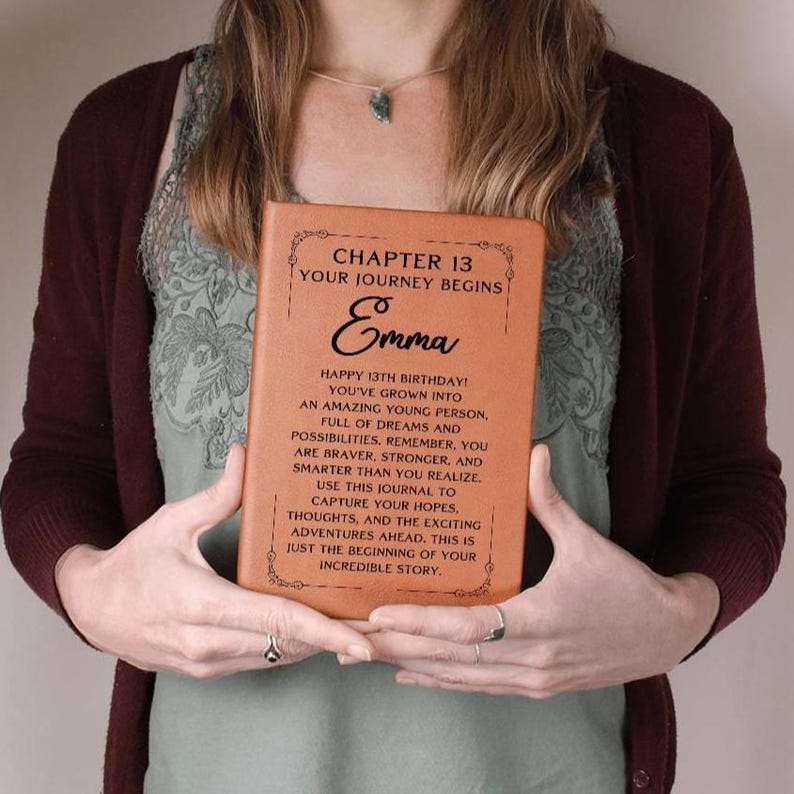 May include: A brown leather journal with the text "Chapter 13 Your Journey Begins" and the name "Emma" in a decorative font. The journal is a gift for a 13th birthday and includes a message about growing up and capturing hopes and dreams.