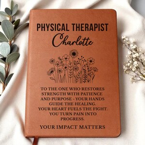 May include: A brown leather-bound journal with the words "PHYSICAL THERAPIST Charlotte" in black lettering. Below the name is a floral design. The journal also includes an inspirational quote about restoring strength and turning pain into progress.