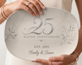 May include: An oval-shaped ceramic platter featuring a light beige background with gray botanical illustrations. The text reads 25 SILVER ANNIVERSARY, EST. 2001, and Emily & Jason, accompanied by a small heart icon centered below the number 25.