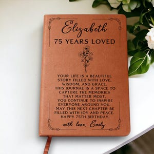 May include: A brown leather journal with the name "Elizabeth" and "75 YEARS LOVED" printed on the cover. The journal includes a floral design and a personalized message. A ribbon bookmark is visible.