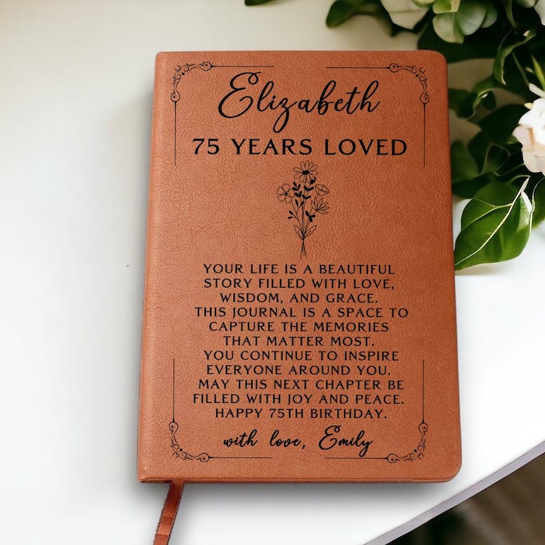 May include: A brown leather journal with the name "Elizabeth" and "75 YEARS LOVED" printed on the cover. The journal includes a floral design and a personalized message. A ribbon bookmark is visible.