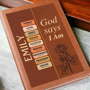 May include: A brown leather-bound journal with the name "EMILY" vertically on the left side. The cover features the text "God says I Am" and a rose illustration. Additional text includes Bible verses and words like "Eternal" and "Loved".