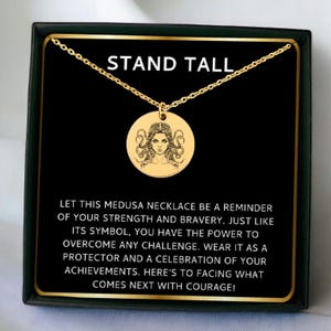 May include: Gold necklace with a round pendant featuring a black and white illustration of Medusa. The necklace comes in a black box with a gold border and the text "STAND TALL" and a message about strength and bravery.