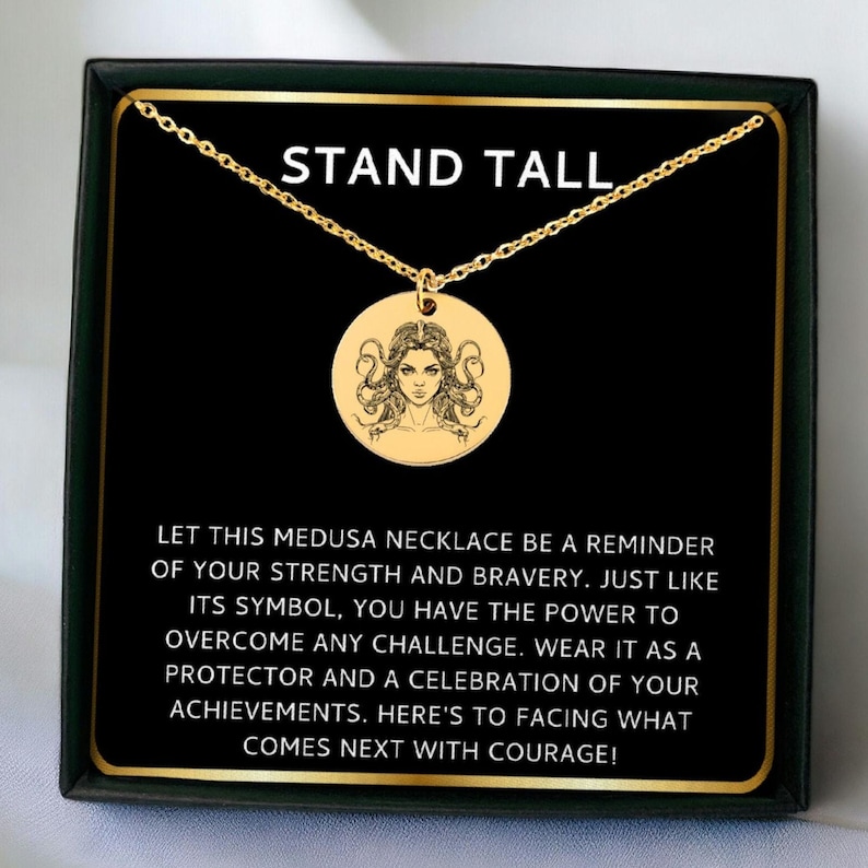 May include: Gold necklace with a round pendant featuring a black and white illustration of Medusa. The necklace comes in a black box with a gold border and the text "STAND TALL" and a message about strength and bravery.