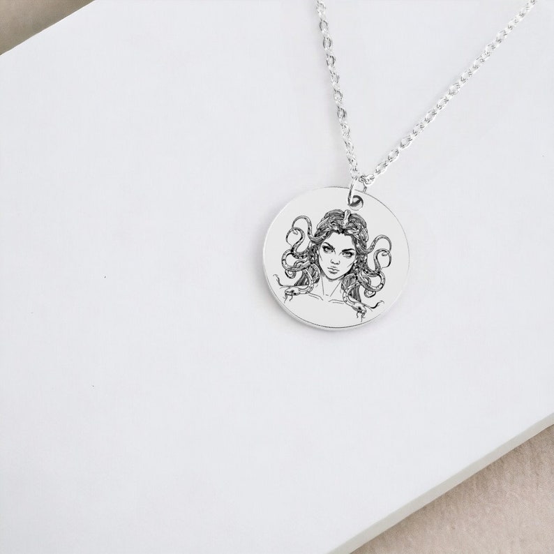 May include: A silver necklace with a round pendant featuring a black and white illustration of Medusa, a Greek mythological figure with snakes for hair.