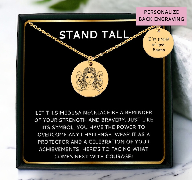 May include: Gold necklace with a round pendant featuring a detailed illustration of Medusa. The pendant is in a black box with the words "Stand Tall" and a message about strength and bravery. The back of the necklace can be personalized with an engraved message.