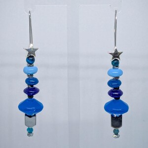 以下が含まれることがあります： 星型のトップと、さまざまな色合いの青いビーズが連なった、ぶら下がりタイプのイヤリングです。イヤリングは銀色の金属仕上げで、下部に幾何学的な形をしています。