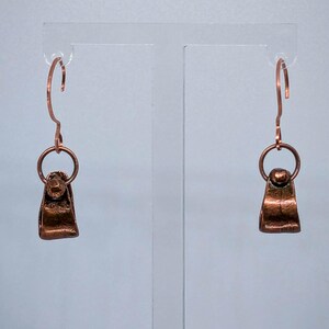 May include: A pair of copper-colored earrings. Each earring features a small, rectangular pendant with a rounded top, connected to a circular ring and a curved ear wire. The earrings are displayed against a light blue background.