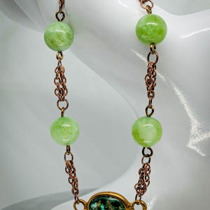 Op de afbeelding: Een ketting met vier lichtgroene, ronde kralen verbonden door koperkleurige schakels. De ketting heeft een ovaalvormige hanger met een groen en goud ontwerp.