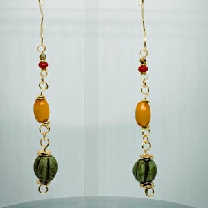 May include: Gold-tone dangle earrings with a unique design. Each earring features a gold hook, small red beads, an amber-colored oval bead, and a green, textured, pumpkin-shaped bead. The earrings are displayed on a clear stand.