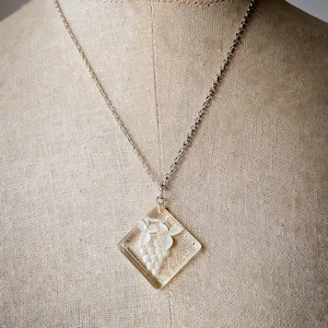 May include: A silver chain necklace with a clear, diamond-shaped pendant. The pendant features a white grape design. The necklace is displayed on a mannequin.