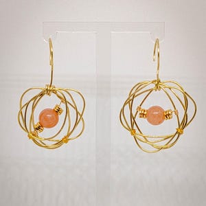 May include: A pair of gold-toned wire-wrapped earrings featuring a spherical peach-colored bead suspended within a cage of concentric circular wire loops. Each earring is attached to a simple gold-toned hook for hanging from the earlobe.
