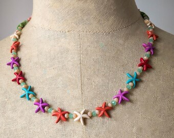 May include: A colorful necklace featuring starfish pendants in red, purple, turquoise, and white, interspersed with small, light green beads. The necklace is displayed on a mannequin, showcasing its design and colors.