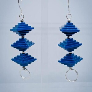 May include: A pair of blue geometric earrings. Each earring features three stacked, square-shaped elements in a vibrant blue hue, connected by silver-colored metal. The earrings dangle from a silver hook and have a small silver ring at the bottom.