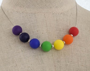 Puede incluir: Un collar de cuentas de colores arcoíris con siete cuentas redondas. Las cuentas son moradas, azul oscuro, azules, verdes, amarillas, naranjas y rojas.
