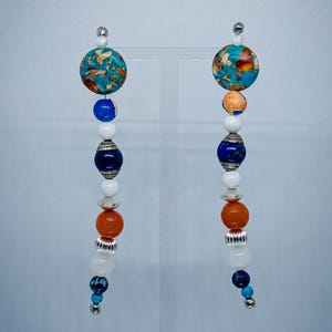 May include: A pair of dangle earrings with a celestial theme. Each earring features a large, round turquoise and orange bead at the top, followed by a series of smaller beads in white, blue, and orange, and silver accents. The earrings are suspended from silver hooks.