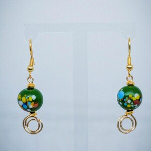 May include: A pair of gold dangle earrings with green glass beads. The beads are covered in colorful confetti-like speckles.