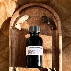 May include: A small, dark brown glass bottle with a black cap and a white label that reads "OldTreePrintShop DO NOT EAT-DRINK BAYOU WATER". The bottle is sitting on a wooden tray with four small, white bone-like objects around it.