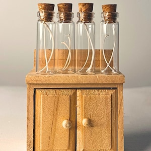 May include: Four clear glass vials with cork stoppers, each containing a single white bone fragment, are arranged in a row on top of a small, light-colored wooden cabinet featuring two doors with round knobs and a flat surface.