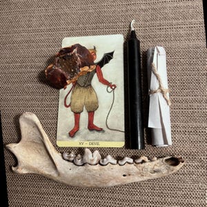 May include: An assortment of occult items, including a tarot card with a devil illustration, a black candle, a rolled parchment tied with twine, and a jawbone. A large, rough-cut crystal is also present.