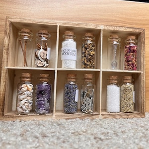 May include: A wooden box with 10 small glass jars with cork tops. Each jar contains different items, such as seashells, lavender, amethyst crystals, and a small silver spoon. The label on one jar reads "Do Not Eat-Drink Moon Dust".