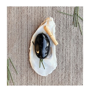 May include: A polished black oval stone rests inside a white oyster shell, accompanied by a small rolled paper and a gold ring. Green sprigs add a natural accent, set against a textured, neutral background.