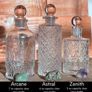 May include: Three clear glass bottles with stoppers, labeled "Arcane", "Astral", and "Zenith". The bottles are approximately 6, 6.5, and 5.5 inches tall with stoppers. Each bottle has a different design and is accompanied by a gemstone.