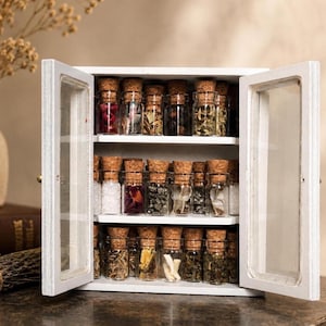 May include: A white wooden cabinet with two hinged glass doors featuring three shelves filled with small clear glass vials. Each vial is sealed with a cork stopper and contains various dried botanical elements, crystals, and white granular substances.
