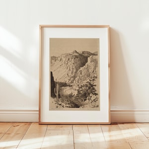 May include: Framed sepia-toned print of a desert canyon landscape. The artwork depicts a towering rock formation, a winding river, and desert vegetation. The print is in a wooden frame and leans against a white wall.