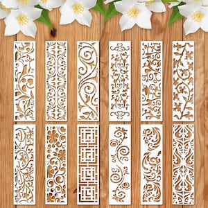 May include: A selection of white, rectangular bookmarks with detailed cut-out patterns. The designs feature floral, geometric, and swirling motifs. The bookmarks are displayed on a wooden surface, with white flowers in the background.