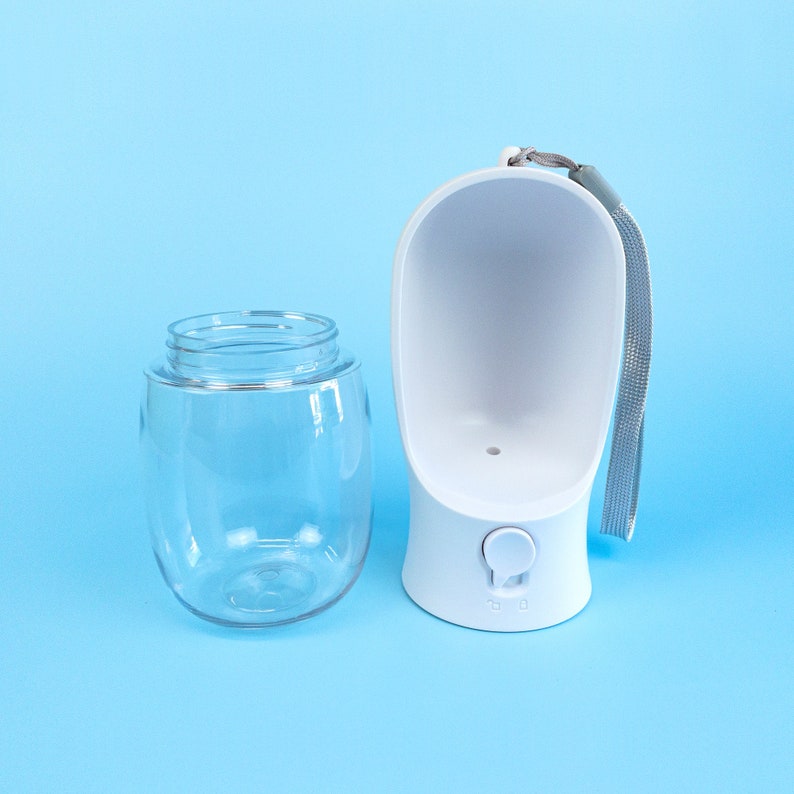 Pu&ograve; includere: Una bottiglia d'acqua per cani in plastica bianca con un serbatoio in plastica trasparente e un cinturino grigio. La bottiglia ha un pulsante per erogare l'acqua.