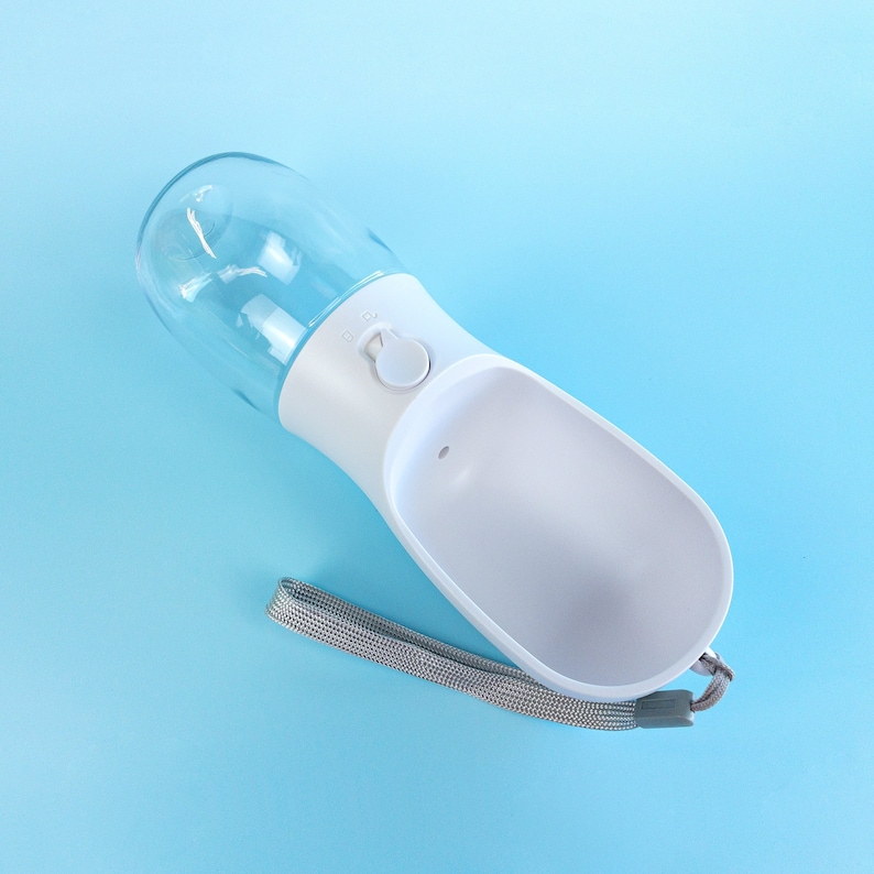 Pu&ograve; includere: Bottiglia d'acqua portatile per animali domestici in plastica bianca e trasparente con una tracolla. La bottiglia ha una ciotola integrata e un pulsante per erogare l'acqua.