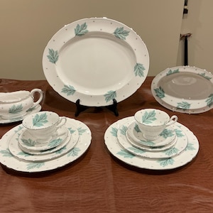 Puede incluir: Un juego de vajilla de porcelana blanca con un patrón de hojas verde azulado y borde plateado. La colección incluye una fuente ovalada grande, un plato ovalado pequeño, una salsera con plato y dos servicios de mesa con taza, platillo y plato.