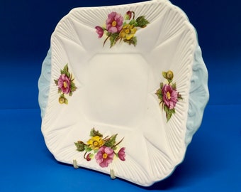 May include: A white square serving dish with a blue rim and floral design. The dish features pink and yellow flowers with green leaves.