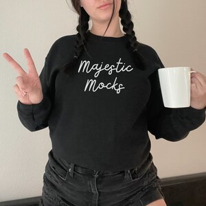 May include: A black sweatshirt with the text "Majestic Mocks" in white script on the front. The person is wearing black jeans and holding a white mug.
