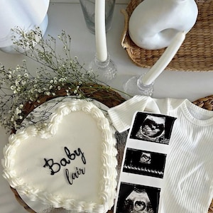 Puede incluir: Un pastel blanco en forma de corazón con las palabras "Baby Lair" escritas en glaseado negro. El pastel está en un plato de mimbre marrón. Hay tres imágenes de ultrasonido en blanco y negro en un mameluco blanco.