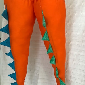May include: Two orange fabric dinosaur leg costumes with blue and green accents. The legs are stuffed and have a series of triangular spikes along the sides. The costume has straps for wearing.