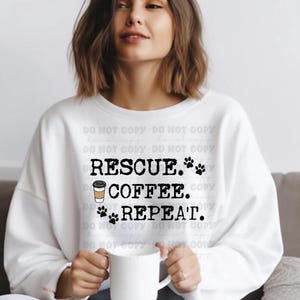 Könnte beinhalten: Weißes Sweatshirt mit dem schwarzen Text "RESCUE. COFFEE. REPEAT." mit Pfotenabdrücken und einer Kaffeetasse. Das Sweatshirt hat einen lässigen Stil.
