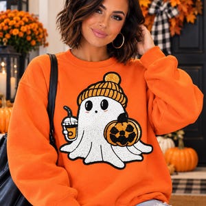 Op de afbeelding: Een oranje sweater met ronde hals en een centrale afbeelding van een witte geest met een gele muts, die een pompoen en een beker vasthoudt. De afbeelding is uitgevoerd in een textuurstijl met zwarte omlijningen.