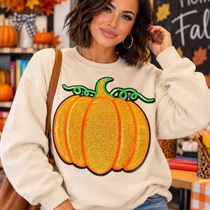 May include: A cream-colored crewneck sweatshirt featuring a large, central graphic of a pumpkin. The pumpkin is filled with bright orange sequins and has a green sequined stem and vine. The design is outlined in a bold black border.
