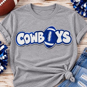 May include: A heather gray short-sleeve t-shirt featuring a large, textured sequin applique across the chest. The design spells out COWBOYS in white and blue sequins, with a blue and white football replacing the letter B.