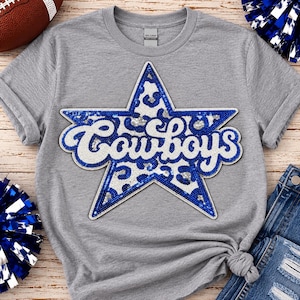 May include: A heather gray t-shirt featuring a large star-shaped patch with a blue and white leopard print pattern. The word Cowboys is written in white script across the center of the star, which is outlined in blue sequins.