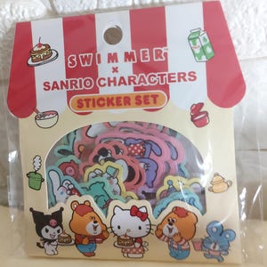 Op de afbeelding: Een sticker set met verschillende Sanrio-personages, waaronder Hello Kitty, in een doorzichtige plastic verpakking. De verpakking heeft een rood-wit gestreept luifelontwerp met de tekst "SWIMMER x SANRIO CHARACTERS STICKER SET". De stickers zijn kleurrijk.