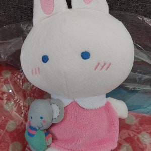 Puede incluir: Un conejito de peluche blanco con un vestido rosa y sosteniendo un ratón de peluche gris. El conejito tiene orejas rosas, ojos azules y una nariz rosa. El ratón tiene pelaje gris, una nariz rosa y una camisa a rayas azul y verde.